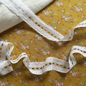 May include: White lace ribbon with a scalloped edge and oval cutouts, displayed on a floral patterned fabric with a yellow background. A rolled piece of white fabric is in the background.
