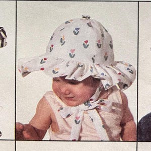 May include: A light-colored baby sun hat with a wide brim and a floral pattern of red, blue, and yellow flowers. The hat has a ruffled edge and is tied with a matching bow. The McCall Pattern Co. All Rights Reserved. Trademarks Reg.