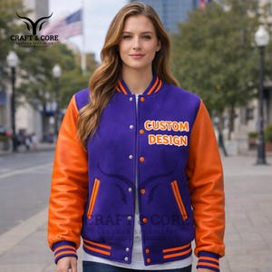 Puede incluir: Chaqueta universitaria morada y naranja con el texto "CUSTOM DESIGN" en el pecho. La chaqueta tiene mangas de cuero naranja y un cuerpo de lana morada. La chaqueta tiene cierre de botones a presión y puños a rayas.
