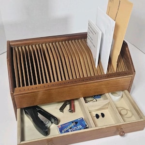 May include: Wooden desk organizer with a drawer and multiple compartments. The top section has vertical dividers for papers, and the drawer contains office supplies like a stapler, staples, tape, and rubber bands. A ruler is etched along the front.