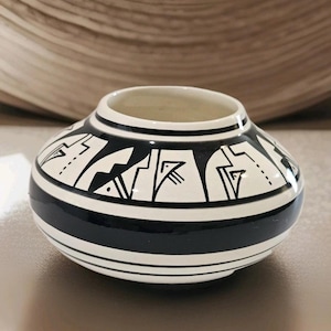 May include: A small, round ceramic vase with a wide opening. The vase is primarily white with a thick black band around the middle and black geometric designs. The vase is approximately 4 inches in diameter and 3 inches tall.