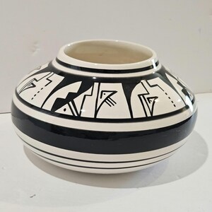 May include: A round, ceramic vase with a wide opening. The vase is primarily white with a bold black stripe around the middle and black geometric designs. The design includes stylized bird and geometric shapes. The vase has a smooth, glossy finish.