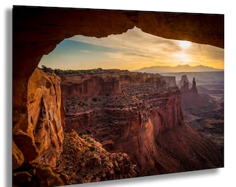 May include: A scenic landscape photograph of a canyon at sunset. The image is framed by a natural arch, with the sun setting over the distant mesas. The canyon walls are a reddish-brown color, and the sky is a mix of orange, yellow, and blue.
