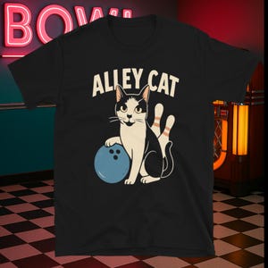 Puede incluir: Camiseta negra con un diseño de gato de dibujos animados. El gato sostiene una bola de bolos azul y está junto a dos bolos. El texto "ALLEY CAT" está en blanco encima del gato.