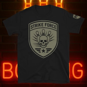 Puede incluir: Camiseta negra con un diseño de escudo. El escudo presenta las palabras "STRIKE FORCE" sobre una calavera y bolos. El diseño es en tonos grises y beige. Una estrella y líneas están en la parte inferior del escudo.