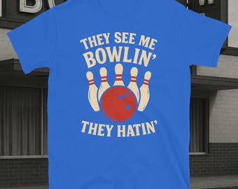 May include: A blue t-shirt featuring the text "THEY SEE ME BOWLIN' THEY HATIN'" in white, along with a graphic of a red bowling ball and pins. The shirt has a crew neck and short sleeves.