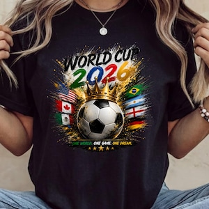 Może przedstawiać: Czarny t-shirt z grafiką piłki nożnej w złotej koronie. Napis głosi WORLD CUP 2026 oraz ONE WORLD. ONE GAME. ONE DREAM. Projekt zawiera kolorowe plamy farby oraz r&oacute;żne flagi państwowe otaczające piłkę.