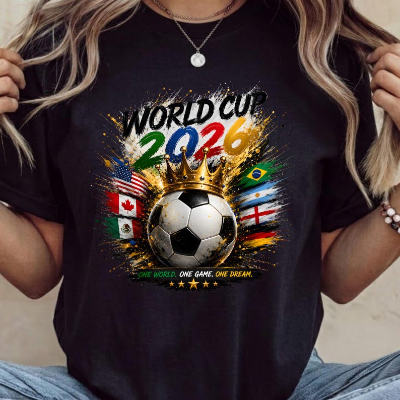 Może przedstawiać: Czarny t-shirt z grafiką piłki nożnej w złotej koronie. Napis głosi WORLD CUP 2026 oraz ONE WORLD. ONE GAME. ONE DREAM. Projekt zawiera kolorowe plamy farby oraz r&oacute;żne flagi państwowe otaczające piłkę.