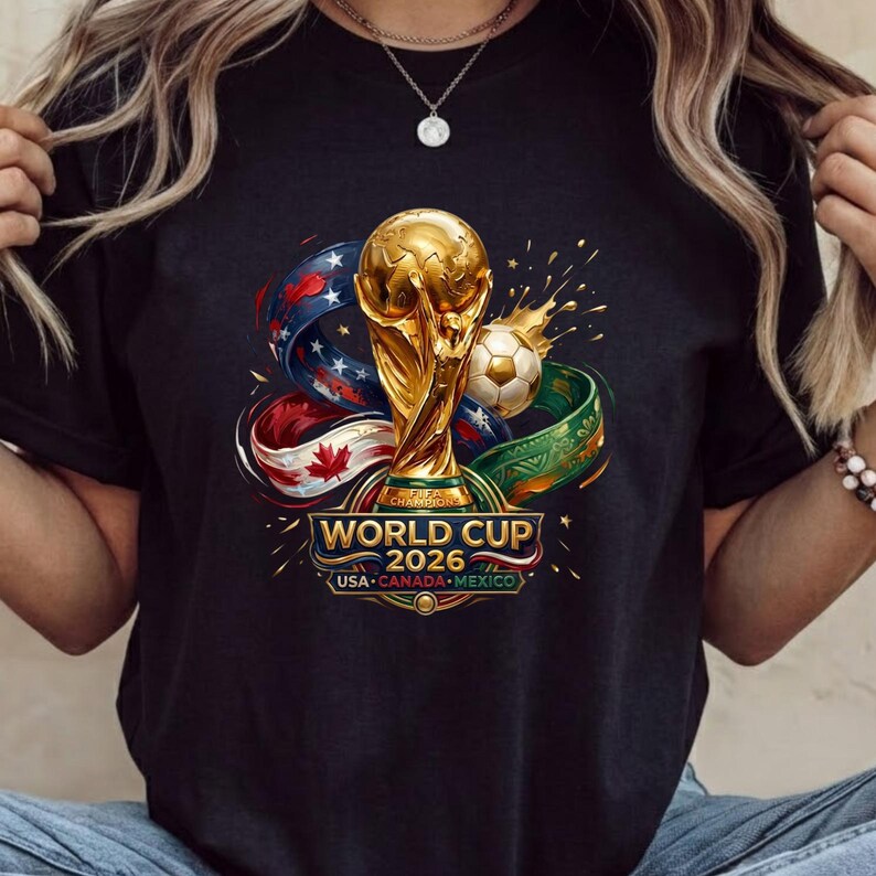 Może przedstawiać: Czarny t-shirt z grafiką złotego trofeum Mistrzostw Świata 2026, piłką nożną i wirującymi wstążkami w barwach flag USA, Kanady i Meksyku. Napis głosi WORLD CUP 2026 USA CANADA MEXICO złotymi i białymi literami na środku koszulki.