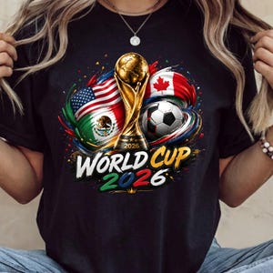 Może przedstawiać: Czarny t-shirt z grafiką złotego trofeum Mistrzostw Świata 2026, piłką nożną oraz flagami Stan&oacute;w Zjednoczonych, Meksyku i Kanady. Wz&oacute;r zawiera kolorowe plamy farby oraz napis World Cup 2026 wykonany pogrubioną czcionką.