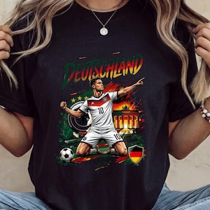 Może przedstawiać: Czarny t-shirt z grafiką piłkarza w białej koszulce z numerem 10, klęczącego na boisku. Wz&oacute;r zawiera napis DEUTSCHLAND, Bramę Brandenburską, piłkę nożną oraz niemiecką flagę umieszczoną w tarczy herbowej.