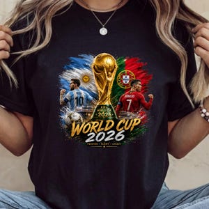 Może przedstawiać: Czarny t-shirt z grafiką trofeum FIFA World Cup 2026 pośrodku, otoczony piłkarzami w koszulkach Argentyny i Portugalii. Tekst głosi FIFA WORLD CUP 2026 PASSION GLORY LEGACY wraz z flagami narodowymi.