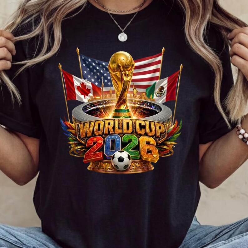 Może przedstawiać: Czarny t-shirt z grafiką przedstawiającą trofeum Mistrzostw Świata FIFA 2026 umieszczone nad piłką nożną. Wz&oacute;r zawiera flagi Kanady, Stan&oacute;w Zjednoczonych i Meksyku oraz napis WORLD CUP 2026 wykonany pogrubionymi, kolorowymi literami.