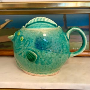 Pode incluir: Uma chaleira de cerâmica turquesa com um design de peixe. A chaleira tem uma tampa, uma alça curva e um bico. O design do peixe inclui barbatanas e olhos. A chaleira tem um acabamento de esmalte craquelado.