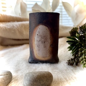 May include: A cylindrical ceramic vessel with a dark, textured charcoal-colored exterior and a prominent, smooth, light-colored oval patch in the center. The surface of the light oval features subtle speckling and a warm, tan-colored border.