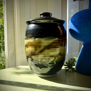 May include: A ceramic jar with a lid, featuring a glossy black glaze on the upper portion and a mottled, earthy blend of olive green, brown, and cream tones on the lower body. The vessel has a rounded silhouette and a smooth, reflective finish.