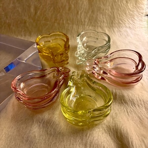 May include: A collection of five small, pear-shaped glass dishes in translucent yellow, clear, and pink hues. Each dish features a smooth, curved rim and a hollow center, arranged in a cluster. The glass material is reflective and glossy.