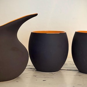 May include: A set of three ceramic vessels. The largest is a dark brown pitcher with an orange rim. Two smaller, rounded black cups with orange interiors are also present. The objects are displayed on a white surface.