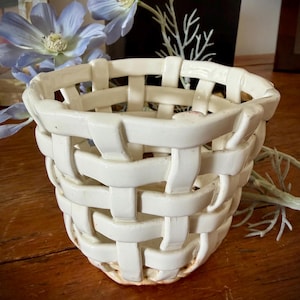 May include: A white ceramic basket with a woven design. The basket is cylindrical with a slightly flared top and sits on a wooden surface. The background includes blue flowers and a dark decorative object.