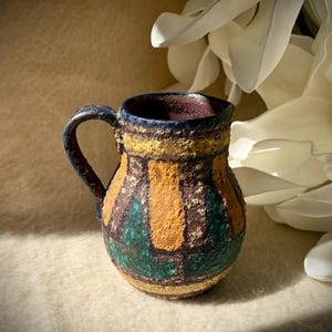May include: A small ceramic pitcher with a textured, rough surface featuring a geometric pattern of mustard yellow, dark teal, and deep brown. The vessel has a rounded body, a single curved handle, and a dark interior, showcasing a rustic finish.