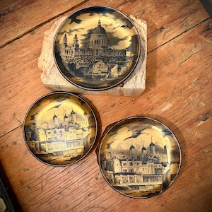 May include: Three decorative plates with gold and black illustrations of buildings. The plates have a gold background with black architectural details. One plate rests on a stone block, while the others sit on a wooden surface.