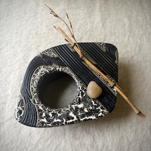 May include: A black and white ceramic object with a central circular opening, featuring a textured crackle pattern and raised linear details. A thin, light brown twig and a small, smooth, tan-colored pebble are placed across the top surface.