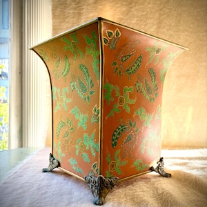 Pode incluir: Um cesto de lixo metálico de formato quadrado com uma base na cor laranja queimado e um padrão floral repetitivo inspirado em paisley em tons de verde e dourado. O recipiente tem uma borda superior alargada e quatro pés metálicos ornamentados.