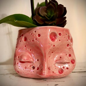 May include: A pink ceramic face planter with a glossy finish, featuring a face design with closed eyes and a textured surface with red accents. The planter holds a succulent plant with green leaves and a dark maroon flower.