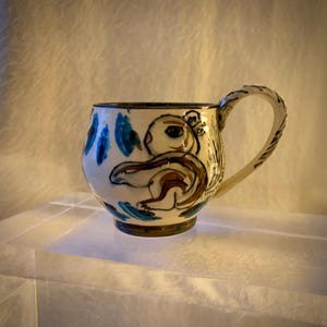 May include: A ceramic mug with a cream-colored base featuring a hand-painted brown squirrel illustration. The design includes blue leaf-like accents around the body and a curved handle with dark brown linear markings along the outer edge.