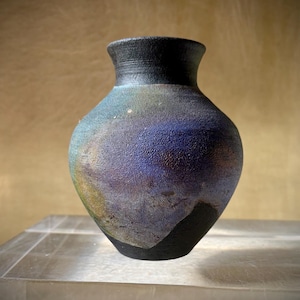 May include: A small ceramic vase with a textured, matte black finish on the neck and base. The rounded body features a mottled, iridescent glaze with blended hues of deep blue, purple, and hints of green, creating a dark, atmospheric appearance.