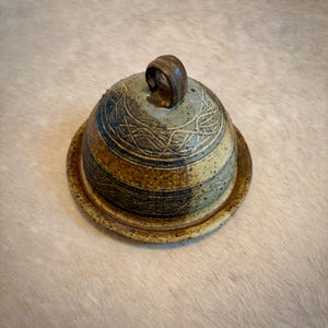 Könnte beinhalten: Eine handgefertigte Butterdose aus Keramik mit Deckel. Die Dose ist rund und hat eine braune, blaue und beige Farbgebung. Der Deckel hat einen Schlaufengriff und geätzte lineare Designs. Die Dose steht auf einem passenden Sockel.