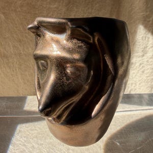 May include: A dark bronze-colored ceramic vase featuring a sculpted, three-dimensional animal head on its front surface. The finish has a metallic, slightly weathered texture with highlights that emphasize the contours of the animal's face.