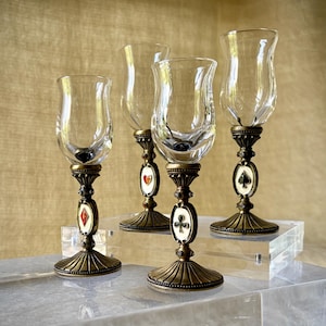 May include: A set of four clear glass goblets with ornate antique bronze-toned metal stems. Each stem features a central oval medallion displaying one of the four playing card suits: heart, diamond, club, and spade, accented with small rhinestones.