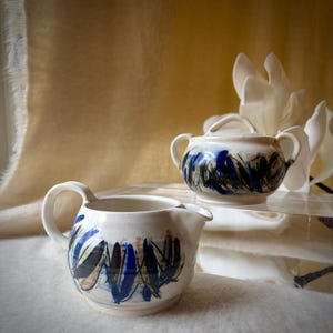 May include: A ceramic creamer and a lidded sugar bowl with two handles, both featuring a speckled off-white glaze decorated with abstract, expressive brushstrokes in shades of deep blue, black, and muted brown around the exterior base.