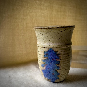 May include: A ceramic tumbler with a speckled cream-colored glaze and a prominent vertical band of textured, deep blue glaze running down the center. The vessel features a slightly flared rim and horizontal ridges along the lower body section.