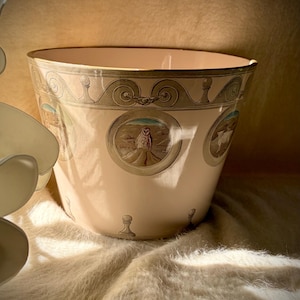 May include: A pale pink ceramic planter featuring a gold-toned rim and a decorative band with circular medallions. Each medallion contains a detailed illustration of an owl in a desert landscape. The base is accented with small, dark, chess-piece motifs.