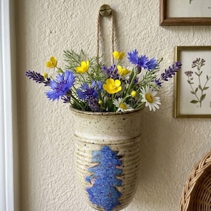 May include: A speckled beige ceramic wall pocket vase with a textured blue vertical accent, hanging from a rope loop. The vase is filled with a bouquet of bright yellow buttercups, vibrant blue cornflowers, purple lavender, and white daisies.