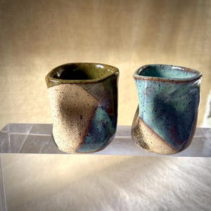 以下が含まれることがあります： 不規則で有機的な形の、手作りの小さな陶器製カップ2つ。左のカップはオリーブグリーンとベージュの斑点のある釉薬、右のカップはターコイズとベージュの釉薬が施されています。どちらも表面に質感があり、斑点模様が見られます。