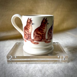 May include: A cream-colored ceramic mug featuring a repeating pattern of orange tabby cats in a sitting position. The mug has a curved handle on the left side and a flared base, showcasing a detailed illustration of the cats around the exterior.