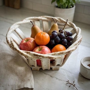 Puede incluir: Un frutero de cerámica blanquecina con frutas frescas. Contiene manzanas rojas, naranjas, uvas oscuras y peras marrones. El frutero tiene un diseño tejido y está sobre una encimera de mármol.