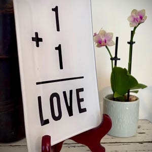 May include: A white rectangular sign with the text "1 + 1 LOVE" in black, displayed on a red stand. The sign is next to a potted orchid with pink and white flowers, creating a decorative display.
