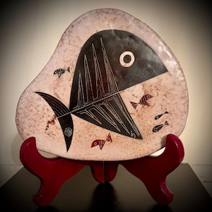 May include: A decorative, abstract ceramic plate featuring a black fish design with white accents and smaller fish illustrations. The plate is displayed on a red stand, showcasing its unique shape and artistic style.
