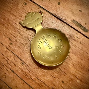 Puede incluir: Una cuchara de latón vintage con diseño de esfera de reloj. La cuchara tiene un mango de tetera y la inscripción "AND ONE FOR ME". Las manecillas del reloj señalan las 11:00. La cuchara descansa sobre una superficie de madera.