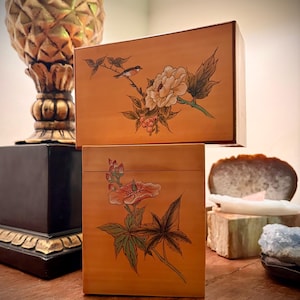 May include: Two wooden boxes with floral and bird illustrations. The boxes are a warm orange color and feature detailed paintings of flowers, leaves, and a small bird. The boxes are stacked on top of each other.