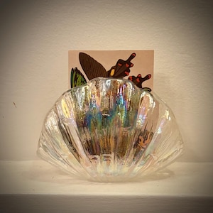 Puede incluir: Un plato iridiscente en forma de concha de vidrio con una ilustración de mariposa. El plato tiene una superficie texturizada y refleja múltiples colores. La ilustración de la mariposa presenta alas marrones, rojas y verdes.