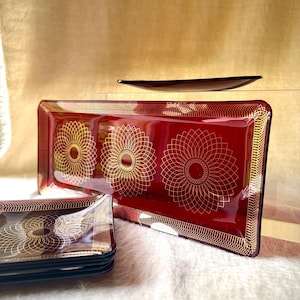 Puede incluir: Una bandeja de servicio rectangular de cristal rojo intenso y una pila de platos cuadrados a juego. Cada pieza presenta un patrón geométrico dorado estilo mandala en la superficie y un borde decorativo de finas líneas doradas.