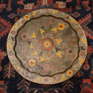 May include: A round brass tray featuring a scalloped edge and an intricate engraved floral pattern. The design includes etched sunflowers and leaves with hand-painted accents in yellow, red, and green, centered around a large circular motif.