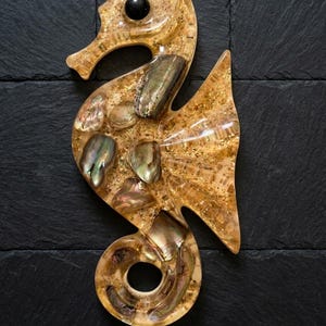 May include: A decorative seahorse figurine in shades of gold and amber, embedded with iridescent shell pieces. The seahorse has a black eye and is set against a dark, textured background. The figurine is approximately 7 inches tall.