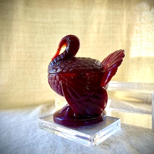 Peut inclure: Un plat à couvercle en verre translucide rouge foncé en forme de dinde. La pièce présente des textures de plumes moulées complexes sur le corps, les ailes et la queue, avec un détail de caroncule distinct sous le cou. La dinde repose sur une base circulaire.