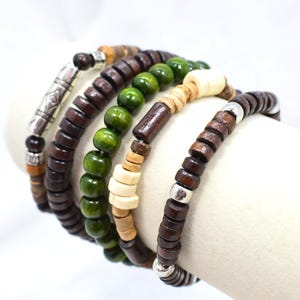 May include: A set of five stacked bracelets featuring dark brown wooden disc beads, round green wooden beads, light tan cylindrical beads, and silver-toned metal accents. The bracelets are arranged in a row, showcasing a variety of textures and colours including earthy brown, vibrant green, and metallic silver elements.